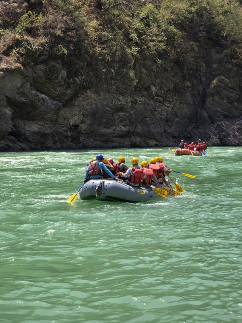 A group of people engaging in outdoor water activity on a raft, paddling along a river with calm, turquoise-green water. The raft is filled with individuals wearing safety gear, including bright yello