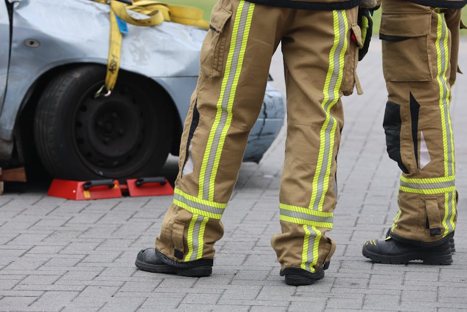 Two emergency responders dressed in tan firefighting turnout gear with yellow reflective stripes are working at the scene of a vehicle incident on a paved street. One of them is standing near a car th